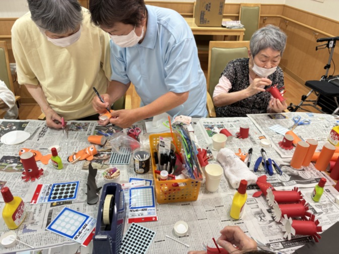 入居者がスタッフと共に工作・手芸活動に取り組む風景。新聞紙を敷いた机での創作レク。