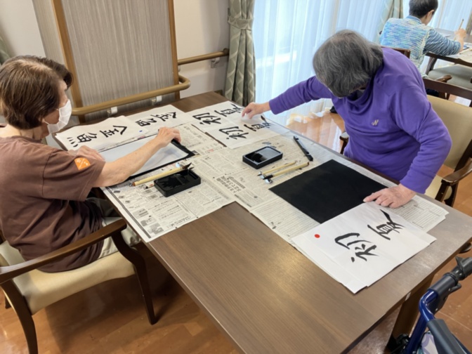 書道レッスン中の入居者と職員。毛筆で文字を書く様子。