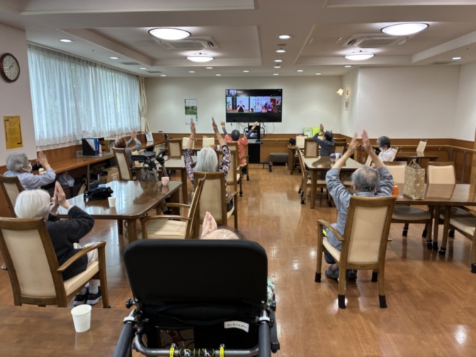 介護施設のリビング・共用部。複数の入居者が座席に配置され、壁掛けテレビで番組を視聴している様子。