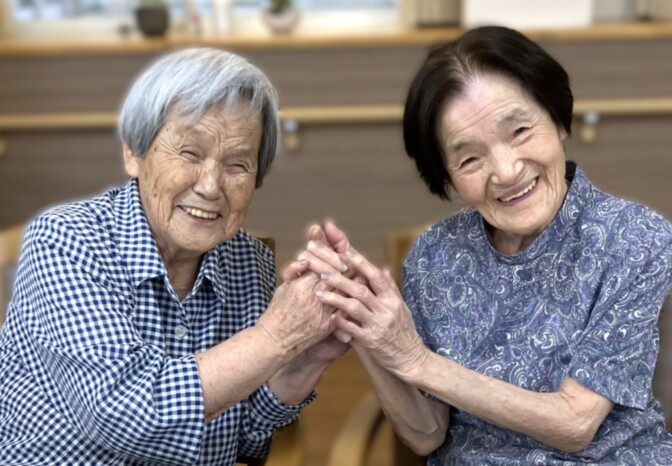 2人の高齢者が手を合わせて笑顔で交流する風景。施設内での温かい日常風景