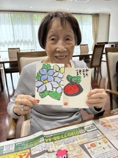 入居者が作成した花と果物の絵を笑顔で掲げている様子