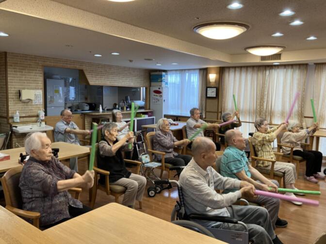 高齢者向けの体操・運動レク活動。複数の入居者が座位で上肢を動かす機能訓練を行っている。
