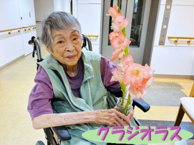 車椅子の入居者が花を持って笑顔で撮影される。施設内での生活風景。