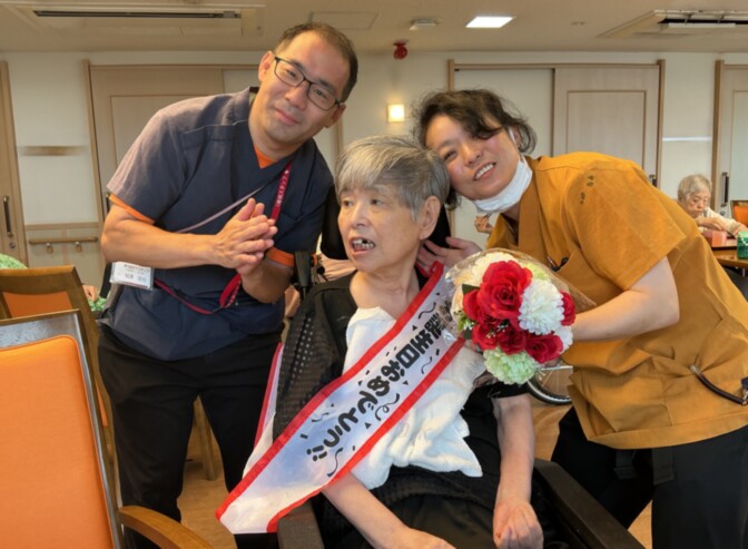 職員と入居者が記念撮花を持って笑顔で写る様子。誕生日や表彰式などのお祝いイベント。