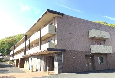 赤茶色レンガ造りの介護施設の外観。モダンな3階建て建物で、バルコニーと駐車スペースがある。