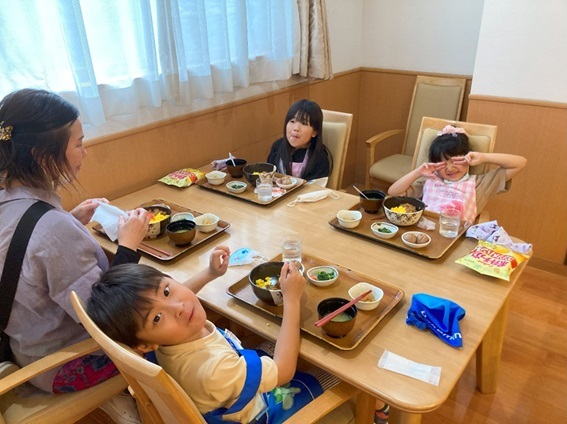 食堂で複数の入居者が食事をしている様子。木製テーブルと椅子、明るい窓からの採光。
